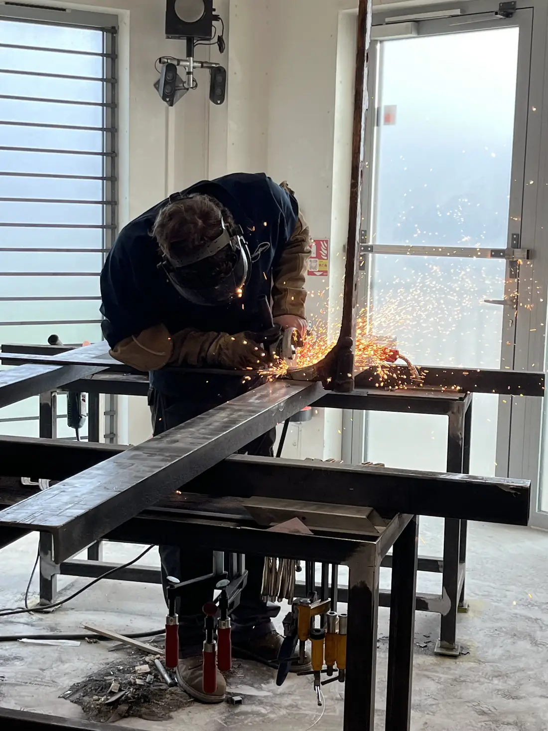 Compagnon meulant un profilé en acier sur un établi de l’atelier, gerbe d’étincelles